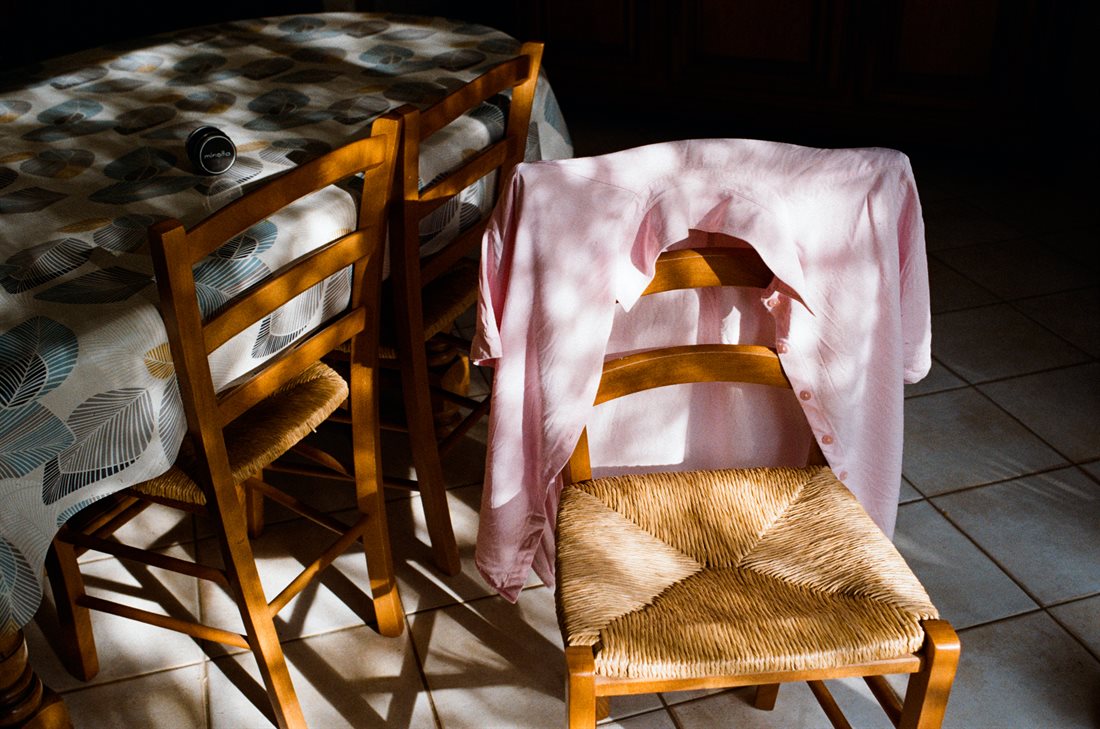 trois chaises assise paille orangées et table avec nappe à dominante bleue, un objectif photo sur la nappe, une chemise rose posée ouverte sur le dossier d'une des chaises